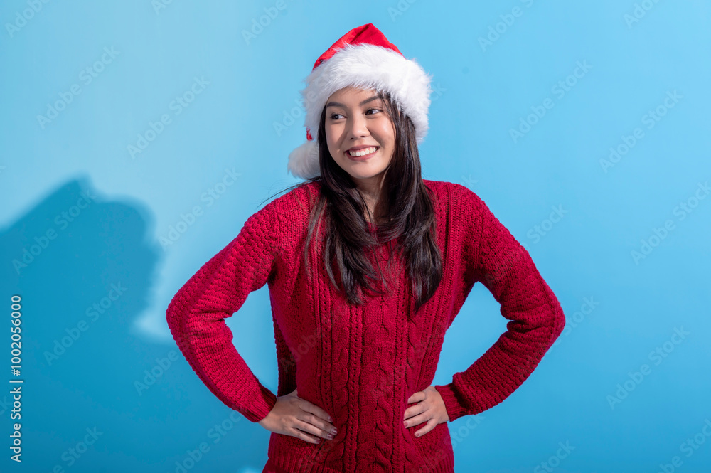 An Asian woman wearing a Santa hat and red knitted sweater stands confidently with her hands on her hips, smiling slightly. The background is a solid light blue, creating a festive holiday theme. An Asian woman wearing a Santa hat and red knitted sweater stands confidently with her hands on her hips, smiling slightly. The background is a solid light blue, creating a festive holiday theme.