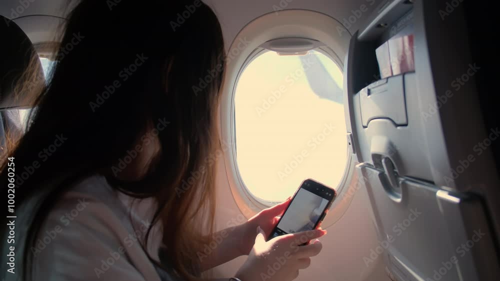 A young woman sits in an airplane and looks through the porthole and takes a photo on the phone