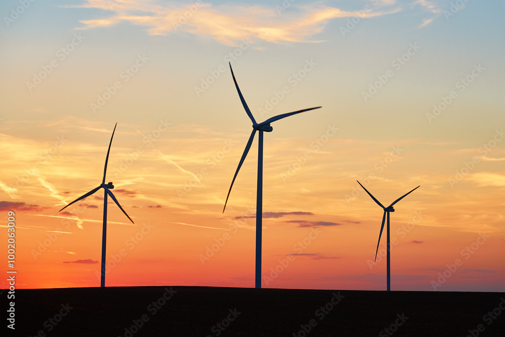 Obraz premium Silhouetted wind turbines in countryside landscape during sunset. Wind power plant generating clean energy. Concept of renewable and sustainable energy