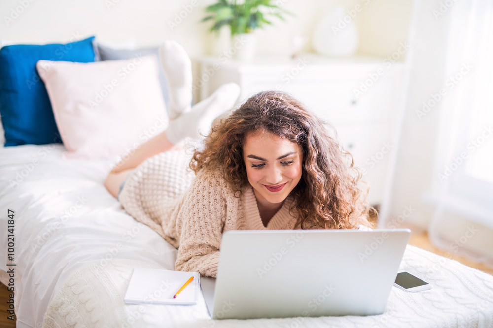 University student attending morning online class on laptop, sitting on bed in dorm room. University student attending morning online class on laptop, sitting on bed in dorm room.