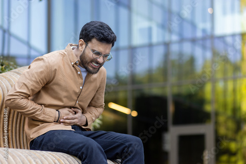 Young man in casual attire sitting on outdoor bench experiencing severe stomach pain. He appears distressed, holding abdomen, conveying discomfort.