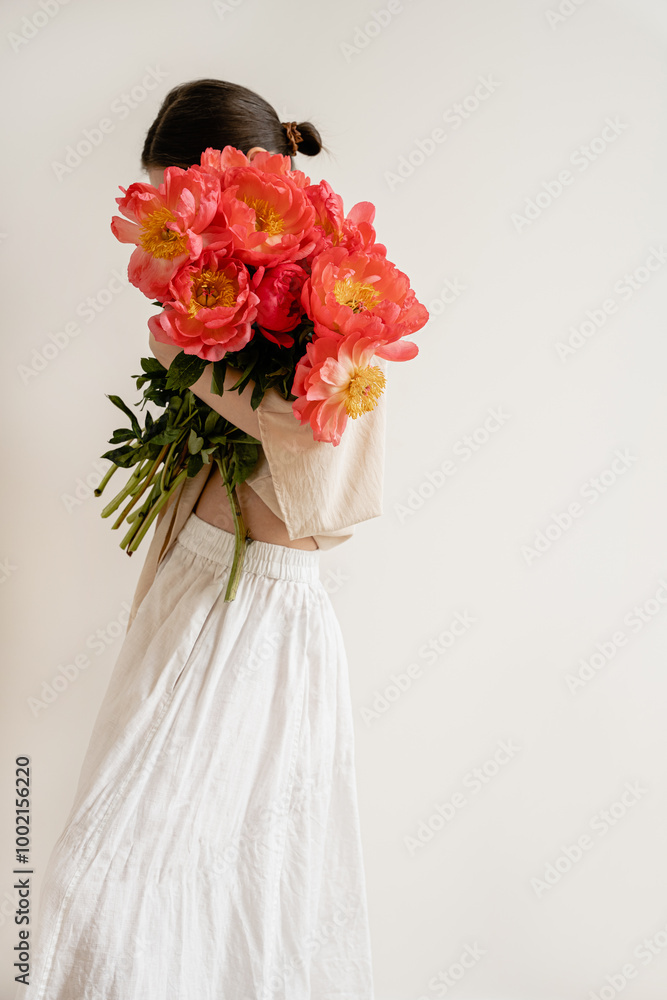 Fototapeta premium Woman in white skirt and blouse embracing large bouquet of bright red peonies. Minimalist style, elegance