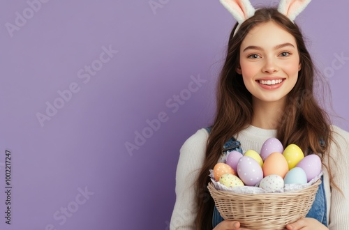 A young girl in bunny ears holds a colorful basket of Easter eggs against a p...