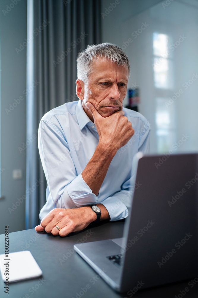 Senior businessman sitting with hand on chin and using laptop at desk in office Senior businessman sitting with hand on chin and using laptop at desk in office