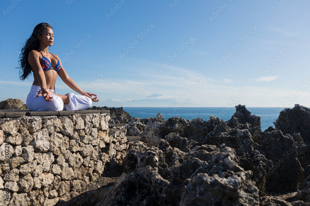 Yoga teacher sitting in lotus position on cliff of Mt Agung under blue sky Yoga teacher sitting in lotus position on cliff of Mt Agung under blue sky
