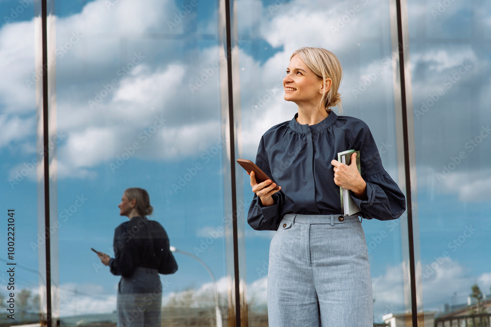 © Westend61 - Smiling businesswoman holding smart phone and diary in front of glass wall