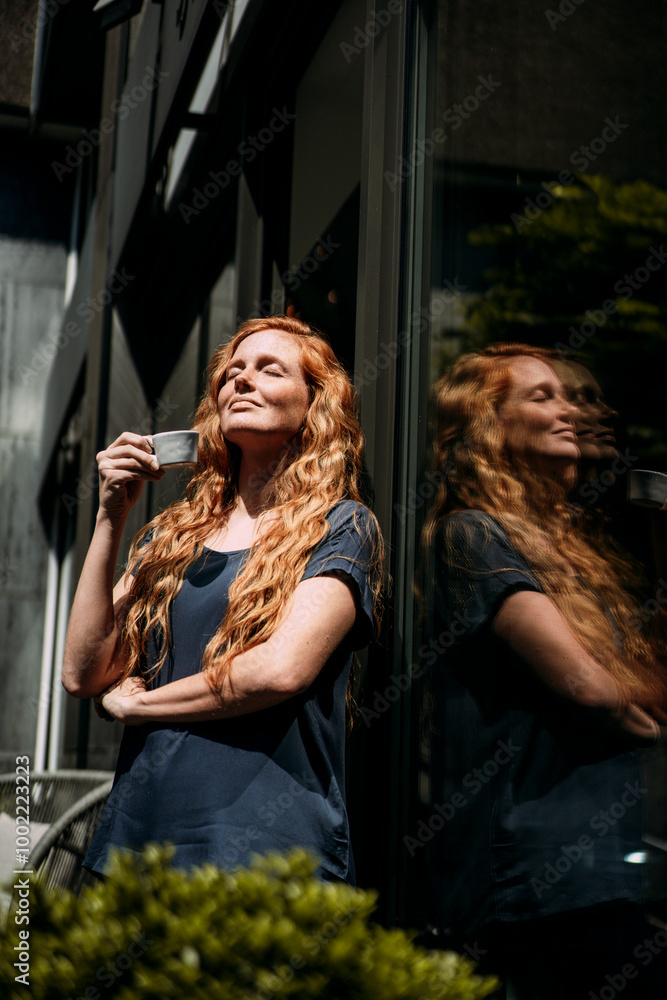 Redhead woman with eyes closed holding tea cup near glass door Redhead woman with eyes closed holding tea cup near glass door