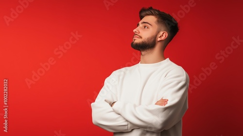 A young man with a beard and crossed arms gazes thoughtfully against a bright red backdrop in an indoor setting.