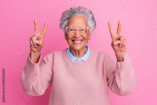 Joyful elderly woman in pink sweater making peace signs against pink background photo