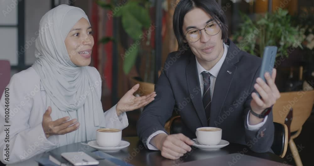 Asian man and Middle Eastern woman in hijab making online video call with smartphone talking to business partners from cafe. Virtual meeting and collaboration concept. Asian man and Middle Eastern woman in hijab making online video call with smartphone talking to business partners from cafe. Virtual meeting and collaboration concept.