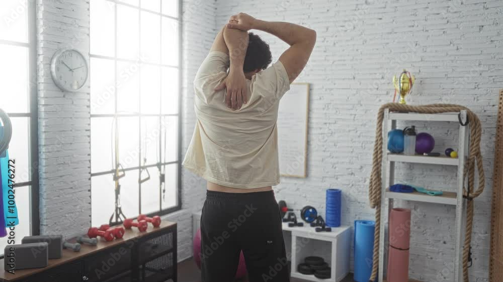 A young hispanic man is stretching indoors in a well-equipped gymnasium with various fitness gear visible, including dumbbells, ropes, and yoga mats. A young hispanic man is stretching indoors in a well-equipped gymnasium with various fitness gear visible, including dumbbells, ropes, and yoga mats.
