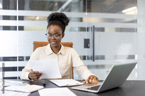 Businesswoman sits at office desk reviewing documents and working on laptop. Engagement in writing and analyzing paperwork, displaying focus and professionalism in corporate environment.