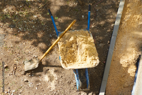 A wheelbarrow filled with sand and a shovel on a construction site in close-up, construction work in the garden, DIY construction, unloading of construction sand, beginning of construction work