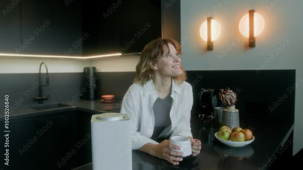 Medium shot of young red-haired Caucasian woman making cup of coffee in kitchen at home, leaning on table, saying command into smart speaker, lights turning on, woman smiling with joy Medium shot of young red-haired Caucasian woman making cup of coffee in kitchen at home, leaning on table, saying command into smart speaker, lights turning on, woman smiling with joy