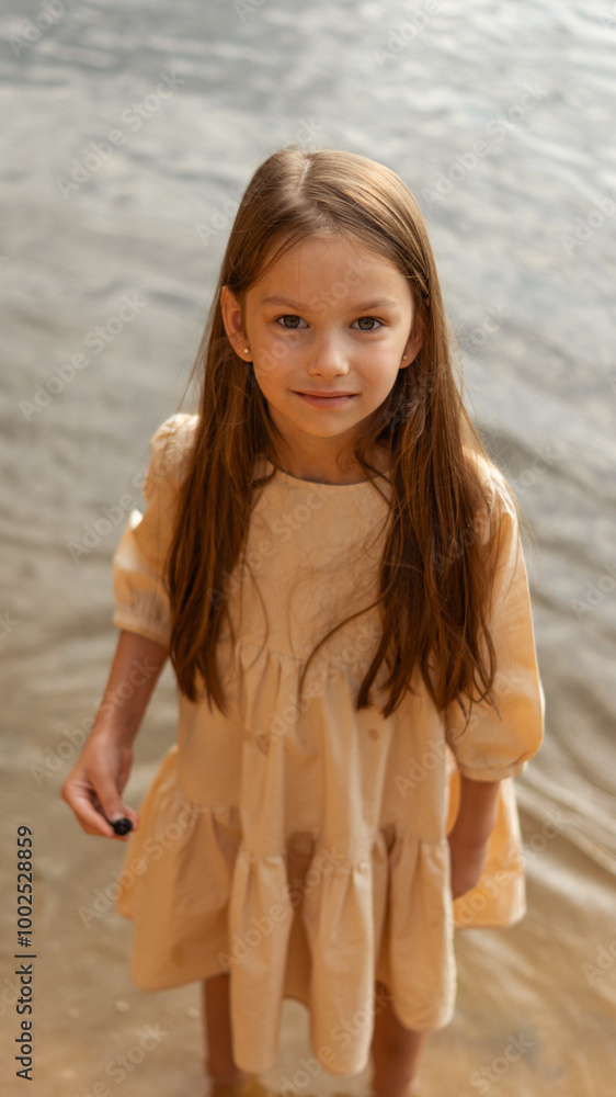 A girl or child with long loose hair in a beige dress stands barefoot in the water on the bank of a river, lake or reservoir, holds a shell or snail or stone in her hands, smiles and looks at the came A girl or child with long loose hair in a beige dress stands barefoot in the water on the bank of a river, lake or reservoir, holds a shell or snail or stone in her hands, smiles and looks at the came