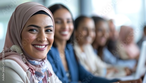 Diverse group of businesspeople, including one Muslim woman wearing a hijab, smiling and sitting at the table with their colleagues in an office setting.