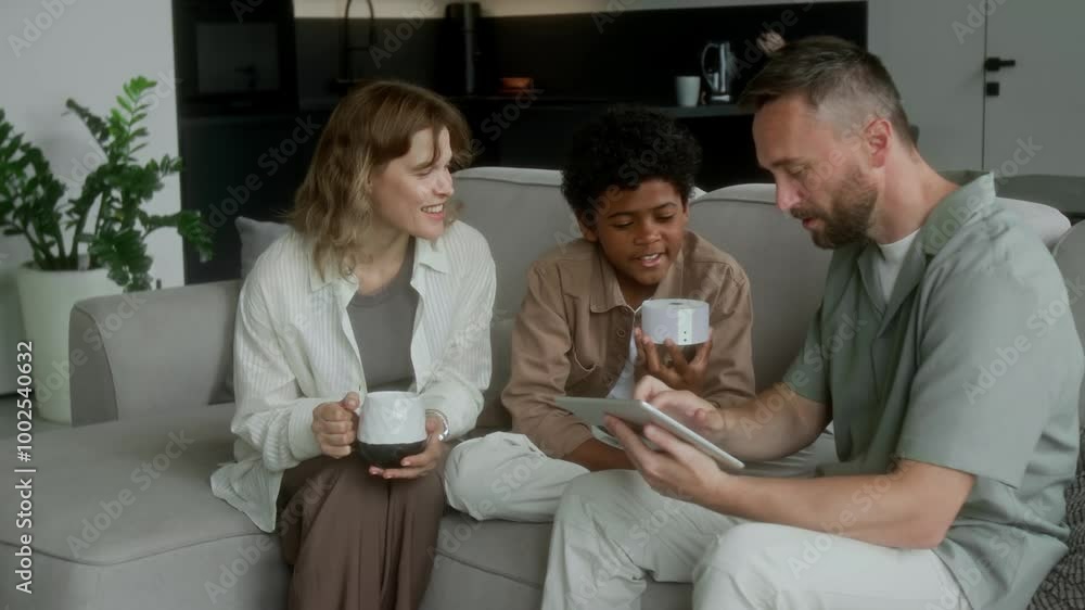 Medium shot of young black boy sitting on couch with foster Caucasian mother and father, saying commands into speaker while learning to use smart home system, adoptive parents laughing Medium shot of young black boy sitting on couch with foster Caucasian mother and father, saying commands into speaker while learning to use smart home system, adoptive parents laughing