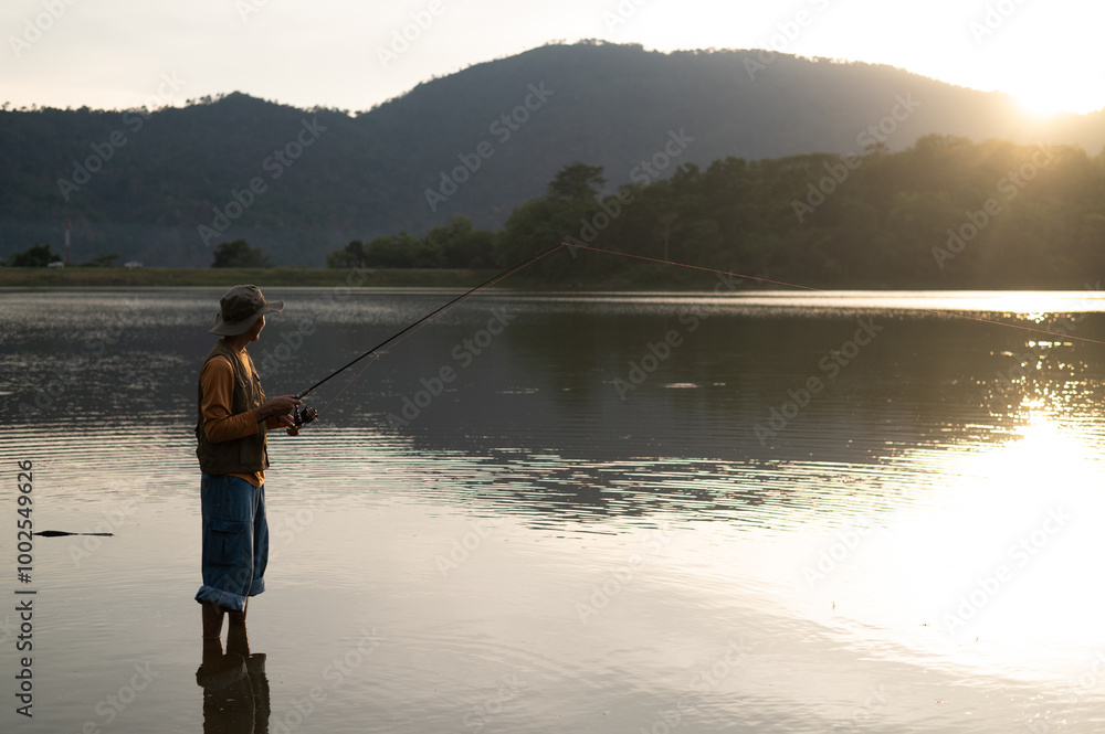 A sunset silhouette of a fisherman fishing with a spinning fishing rod in hand, close-up on the bank of a river or lake. with the setting sun contrasting with the natural forest in the background A sunset silhouette of a fisherman fishing with a spinning fishing rod in hand, close-up on the bank of a river or lake. with the setting sun contrasting with the natural forest in the background