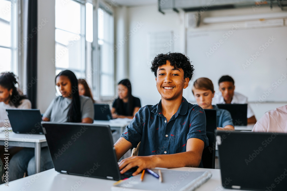 Portrait of smiling curly hair teenage boy sitting with laptop near desk in classroom at school Portrait of smiling curly hair teenage boy sitting with laptop near desk in classroom at school