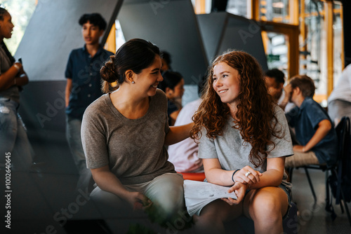 Happy female teacher assisting teenage girl holding book while sitting in cafeteria at junior high school