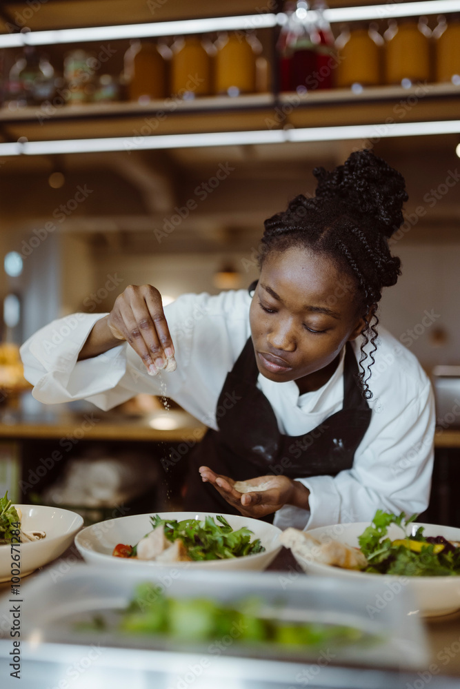 Focused young female chef sprinkling salt on salad while bending in commercial kitchen at restaurant Focused young female chef sprinkling salt on salad while bending in commercial kitchen at restaurant