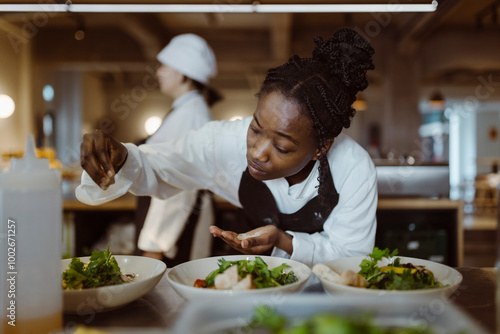 Focused young female chef sprinkling salt on salad while bending in commercial kitchen photo