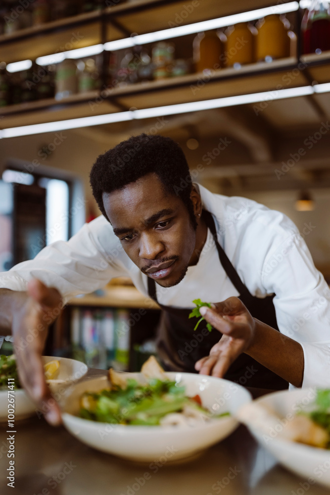 Focused male chef garnishing salad bowl while bending in commercial kitchen Focused male chef garnishing salad bowl while bending in commercial kitchen