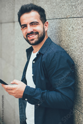 Half length portrait of happy male blogger with cellphone gadegt in hand standing at city area and using public internet connection for cell smartphone device, millennial person with mobile phone photo