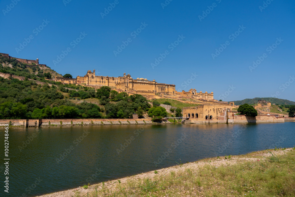 the majestic Amber Fort in Jaipur, Rajasthan, overlooking Maota Lake. The fort’s golden-yellow sandstone facade stands out against the lush greenery and clear blue sky. the majestic Amber Fort in Jaipur, Rajasthan, overlooking Maota Lake. The fort’s golden-yellow sandstone facade stands out against the lush greenery and clear blue sky.