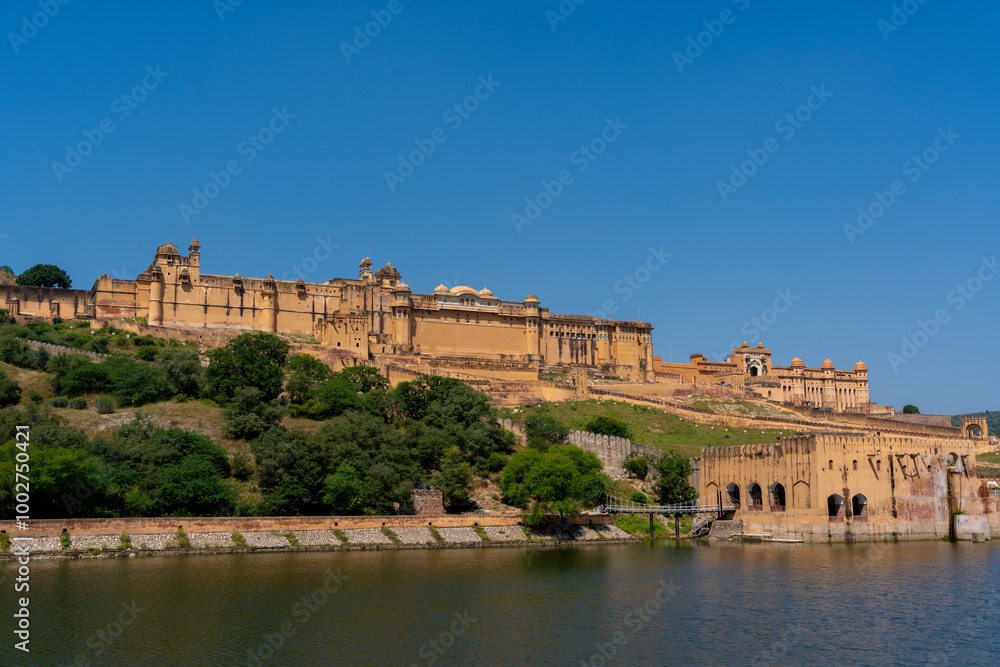 the majestic Amber Fort in Jaipur, Rajasthan, overlooking Maota Lake. The fort’s golden-yellow sandstone facade stands out against the lush greenery and clear blue sky. the majestic Amber Fort in Jaipur, Rajasthan, overlooking Maota Lake. The fort’s golden-yellow sandstone facade stands out against the lush greenery and clear blue sky.