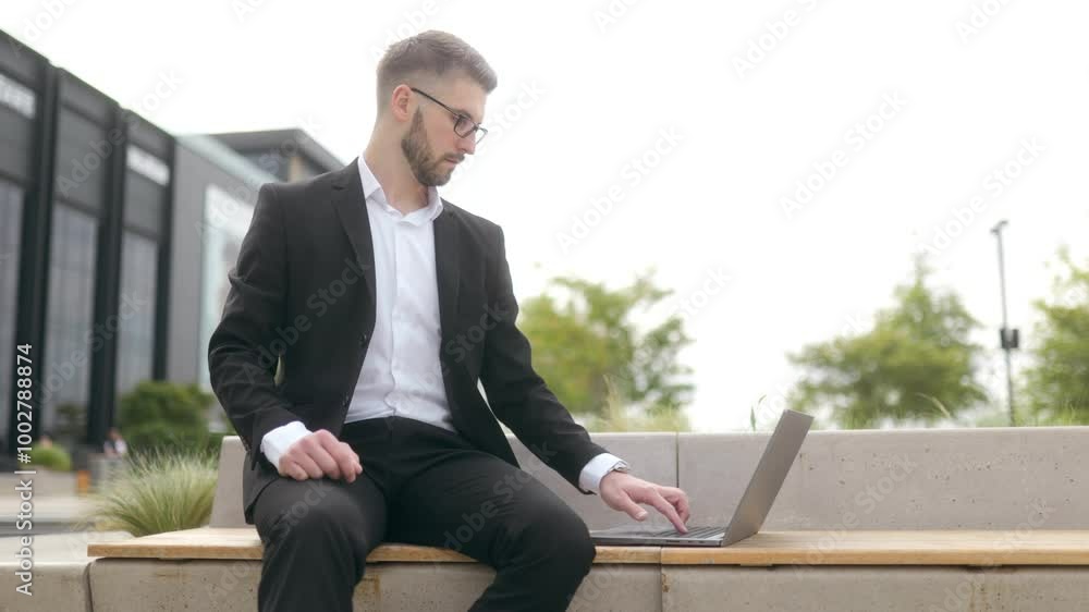 Focused Young Businessman Working on Laptop Outdoors in Modern Urban District Focused Young Businessman Working on Laptop Outdoors in Modern Urban District