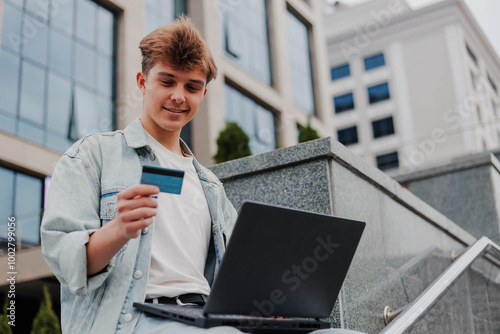 Happy young man using laptop and credit card for online shopping outdoors on a background of corporate building.