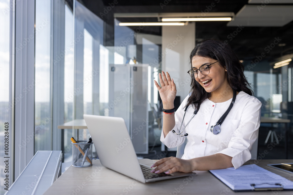 Female doctor engaging in virtual consultation, smiling waving at laptop. Wears stethoscope, suggesting medical context. Office space creates professional atmosphere. Concept of remote healthcare Female doctor engaging in virtual consultation, smiling waving at laptop. Wears stethoscope, suggesting medical context. Office space creates professional atmosphere. Concept of remote healthcare