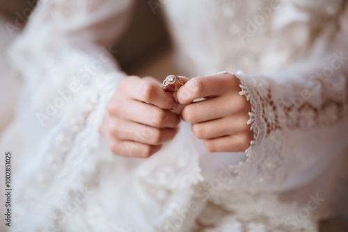 A woman is wearing a white dress and holding a ring in her hand. The dress is white and has lace detailing. The woman is getting ready for a special occasion, possibly a wedding or a formal event photo