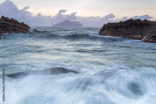 At dusk. Gentle waves lap against smooth, rounded rocks in a tranquil cove. The sky is painted in soft hues of pink and purple, reflecting on the calm water. Toucheng, Yilan, Taiwan.