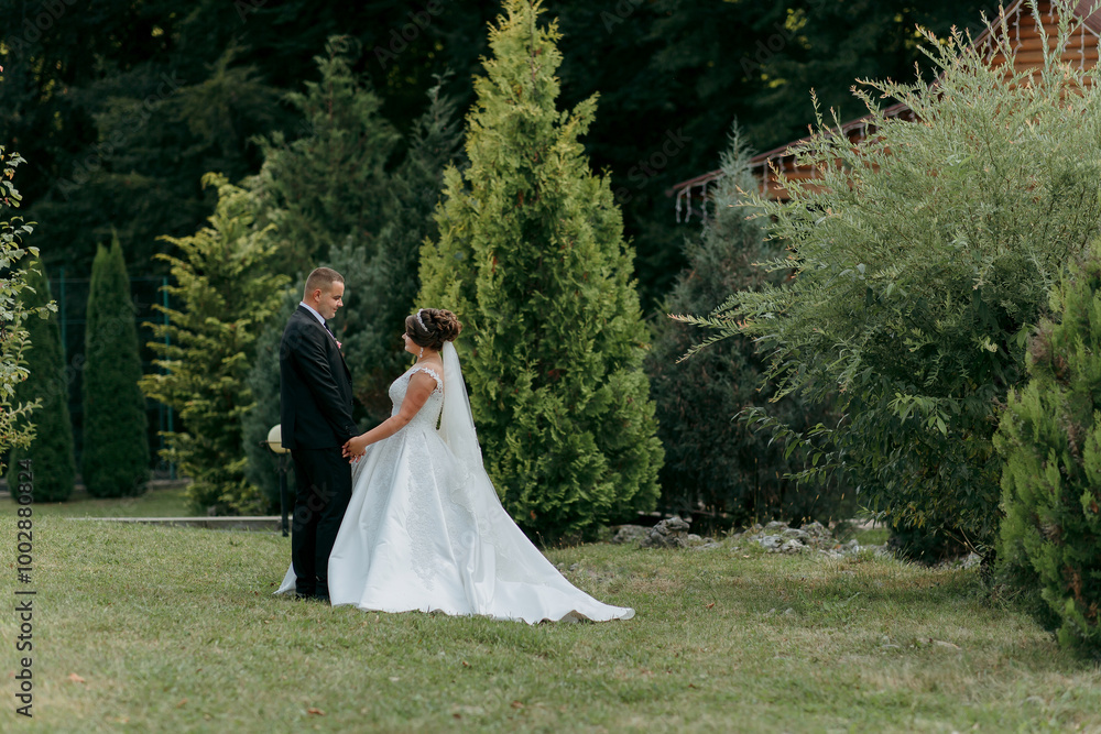 A bride and groom stand in a lush green field, surrounded by trees. The bride is wearing a white dress and the groom is wearing a suit. They are holding hands and smiling at the camera A bride and groom stand in a lush green field, surrounded by trees. The bride is wearing a white dress and the groom is wearing a suit. They are holding hands and smiling at the camera