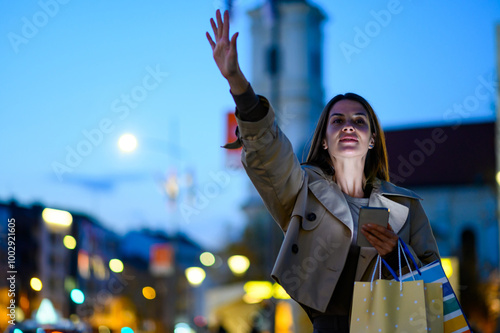 Woman hailing taxi cab in city at night holding shopping bags photo