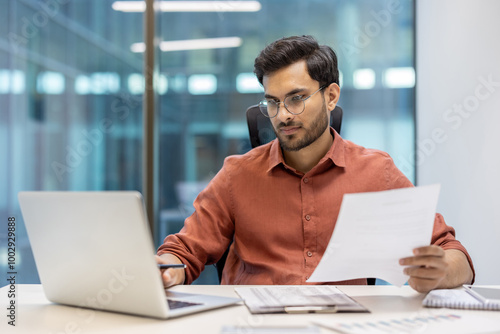 Confident man in glasses working with laptop and holding documents in office. Focused on screen and paperwork, demonstrating concentration and professionalism in business environment.