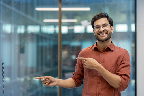 Smiling young man in glasses and casual attire pointing confidently in office setting. Represents positivity, confidence, and professional demeanor in modern workspace.