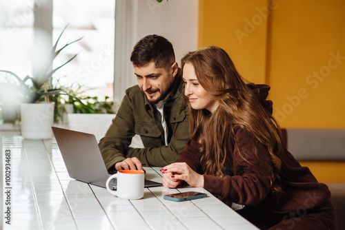 A happy young couple chatting using a laptop on social media while having breakfast in the kitchen. Man and woman looking at computer, watching videos, browsing or shopping in e-commerce stores.