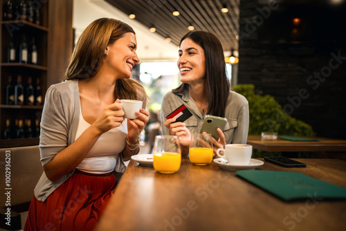 Two girl best friends met at the cafe to shop online together. They are using mobile phone and credit card. Friends hang out and drink coffee and orange juice.