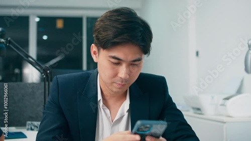 Smilling Asian businessman using smartphone working in office. Attractive Asian male manager working on mobile phone deivce checking email, social media, browsing internet, texting a message. photo