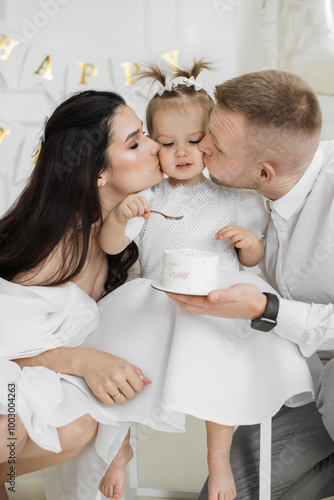 Young Caucasian parents celebrate daughters second birthday. Mother and father kiss girl holding cake with excitement and joy. Warm family moment filled with happiness. photo