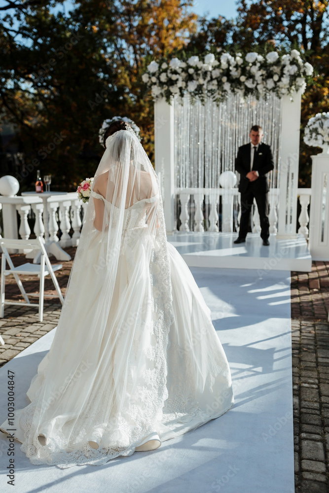 Fototapeta premium A bride is walking down a white carpeted aisle with a groom waiting for her. The bride is wearing a white dress and a veil. The groom is wearing a black suit. The scene is set in a wedding ceremony