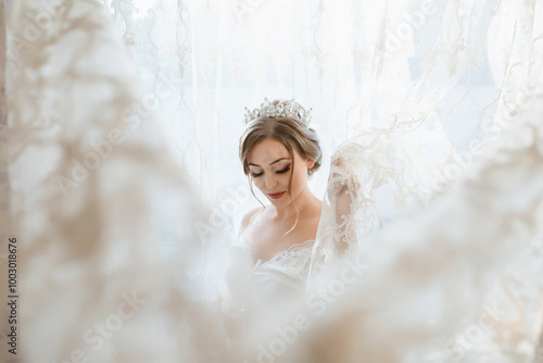 A woman in a white dress is standing in front of a curtain. She is wearing a tiara and has her hair in a bun