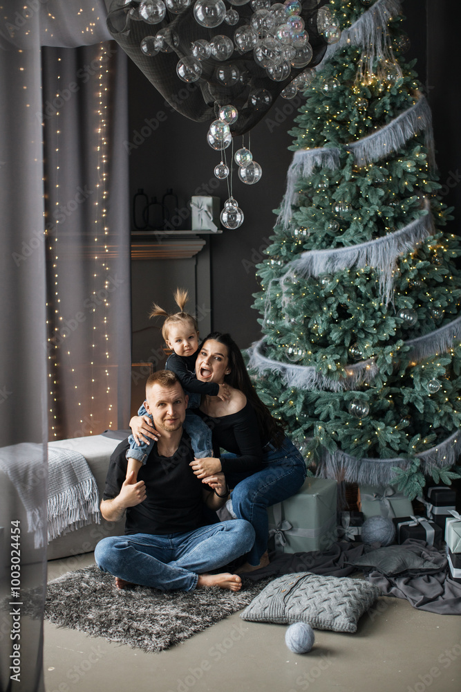 Naklejka premium Cheerful Caucasian family with parents and young daughter enjoying Christmas at home. Cozy living room setting with decorated Christmas tree, presents, and festive lights.