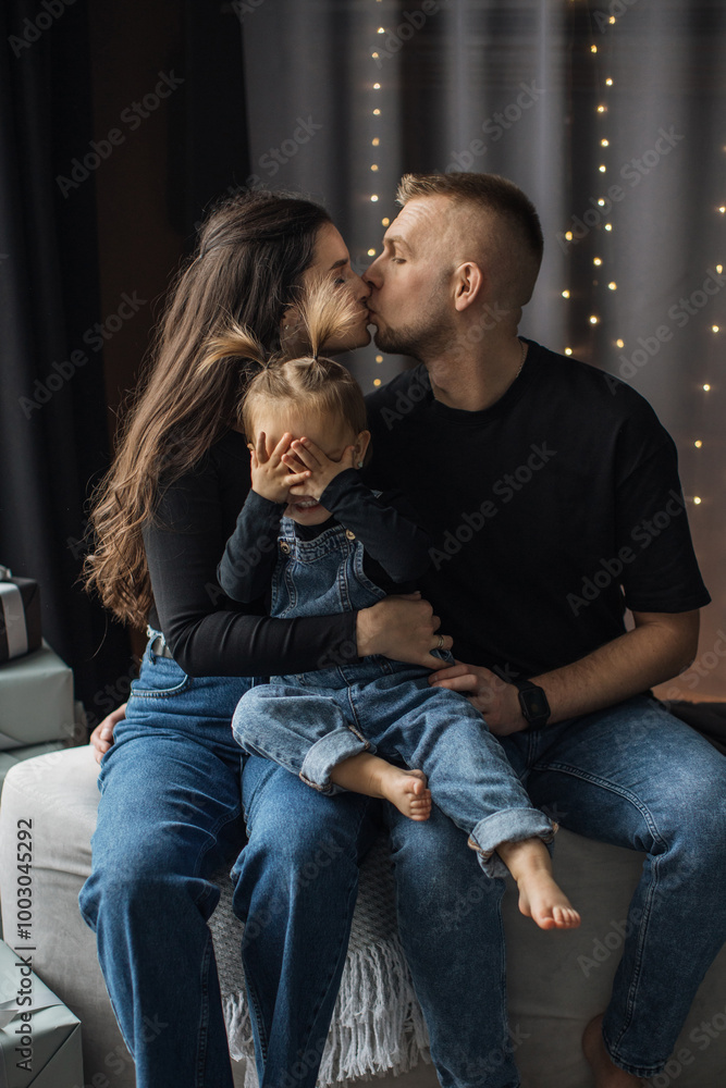 Naklejka premium Caucasian family capturing intimate moment with young parents kissing while child playfully covers eyes. Parents wear casual clothing sitting in cozy home setting.