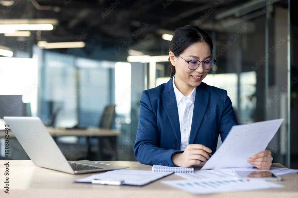 Asian businesswoman analyzes paperwork while working in contemporary office space. Professional woman in glasses reviewing documents beside laptop showcasing business focus and confidence. Asian businesswoman analyzes paperwork while working in contemporary office space. Professional woman in glasses reviewing documents beside laptop showcasing business focus and confidence.