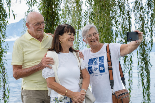 Senior friends walking outdoors along Lake Geneva  take a selfie with cell phone under a weeping willow tree. Smiling senior man and two women enjoying freedom and vacation or retirement #1003100030