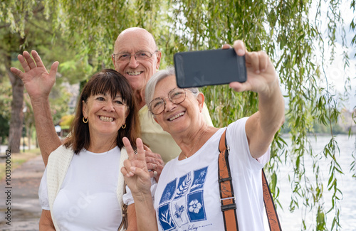 Senior friends walking outdoors along Lake Geneva  take a selfie with cell phone under a weeping willow tree. Smiling senior man and two women enjoying freedom and vacation or retirement #1003100062